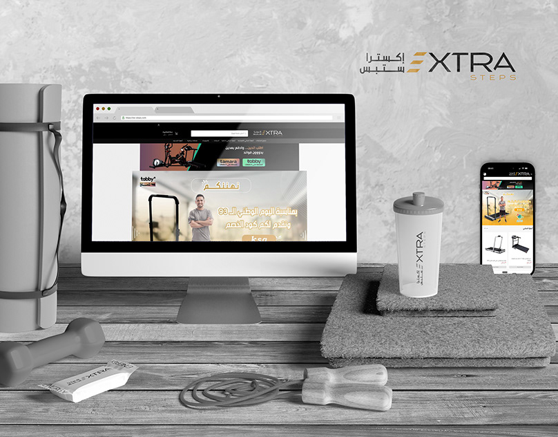 Extra Steps fitness store website shown on a desktop monitor and smartphone beside gym accessories
