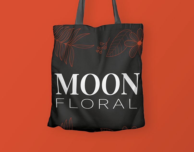 Black canvas tote bag printed with the Moon Floral logo and floral line art on a red-orange background